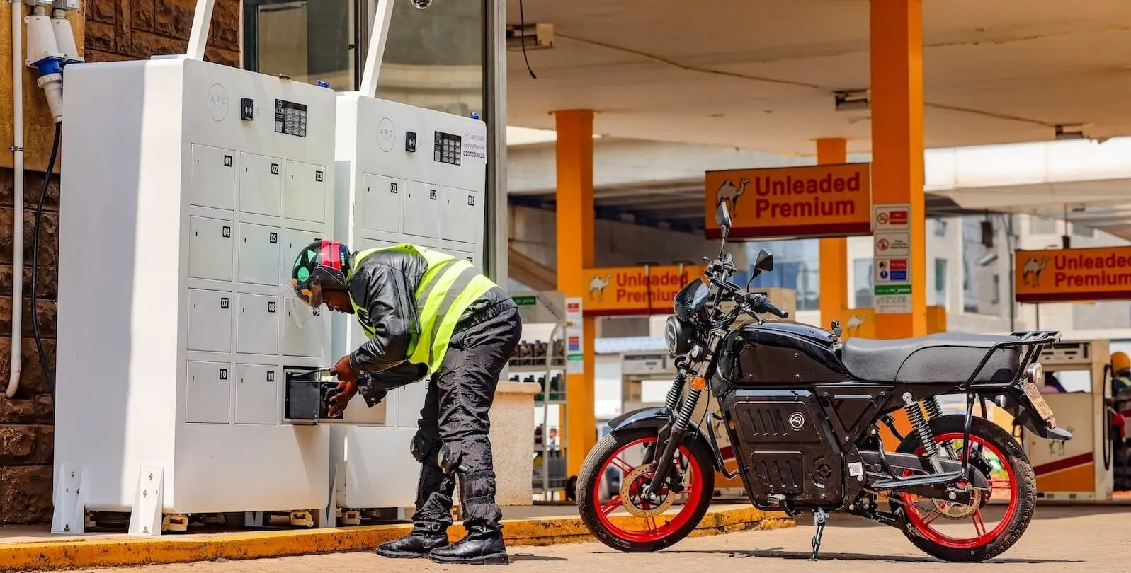A rider swaps a battery at an ARC Ride station next to an electric motorcycle at a petrol forecourt