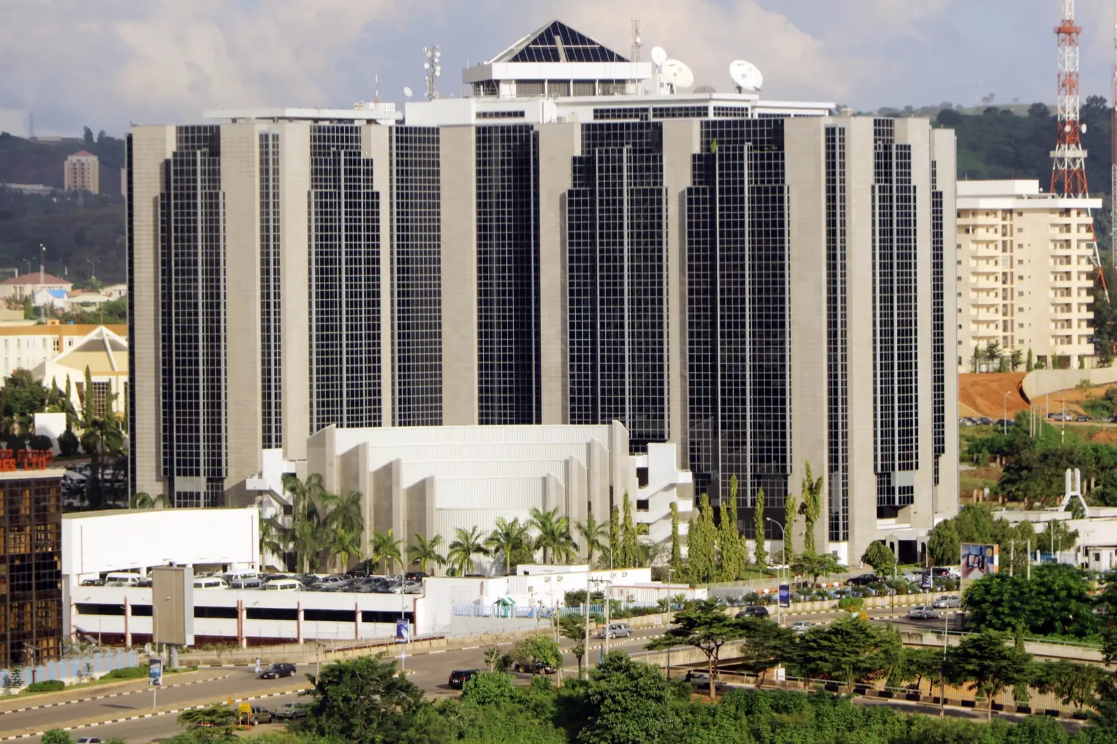 The Central Bank of Nigeria's headquarters in Abuja, a tall grey and black glass tower behind palm trees and a highway