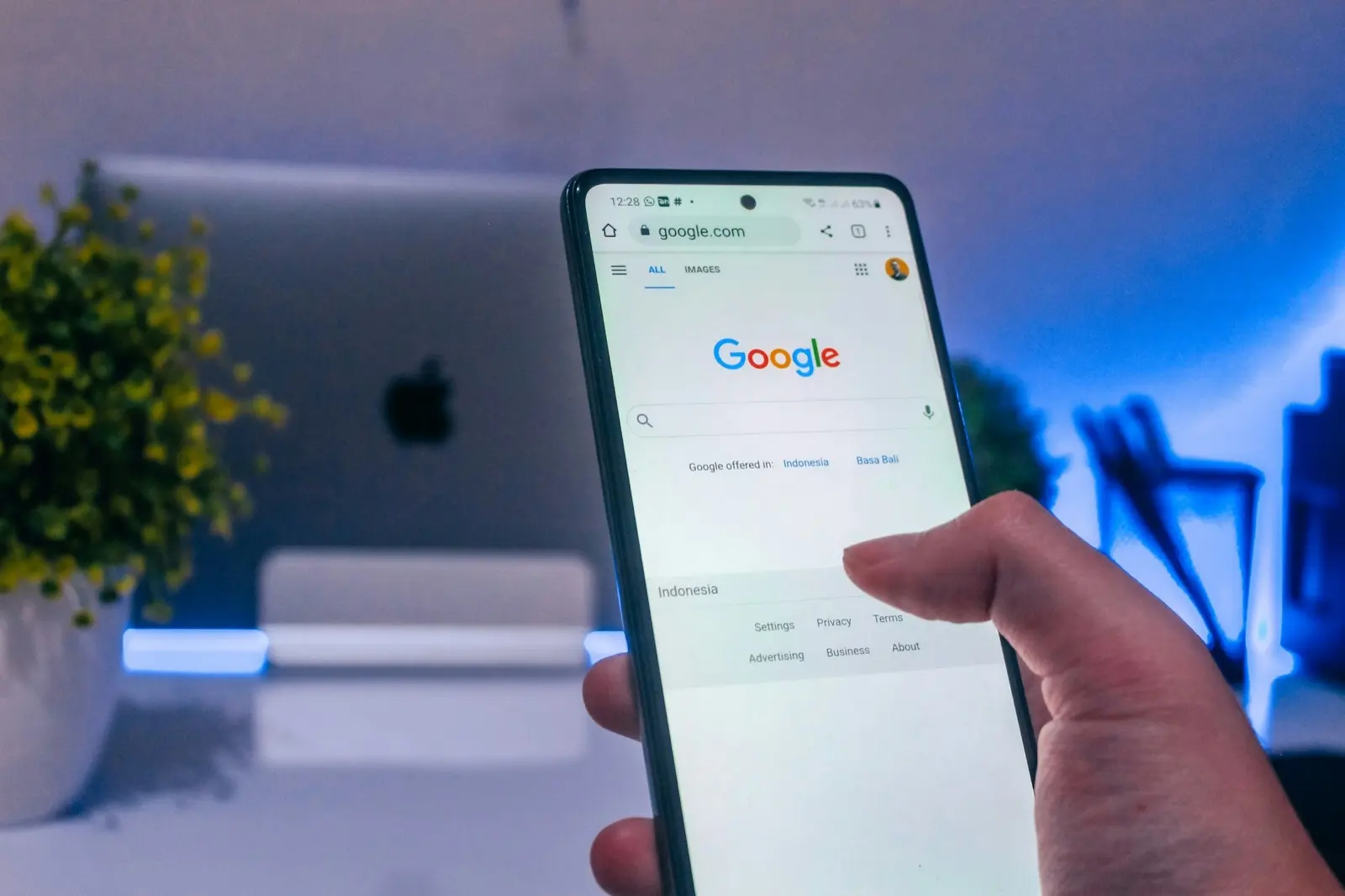 A hand holds a smartphone showing the Google search page in front of a desktop computer