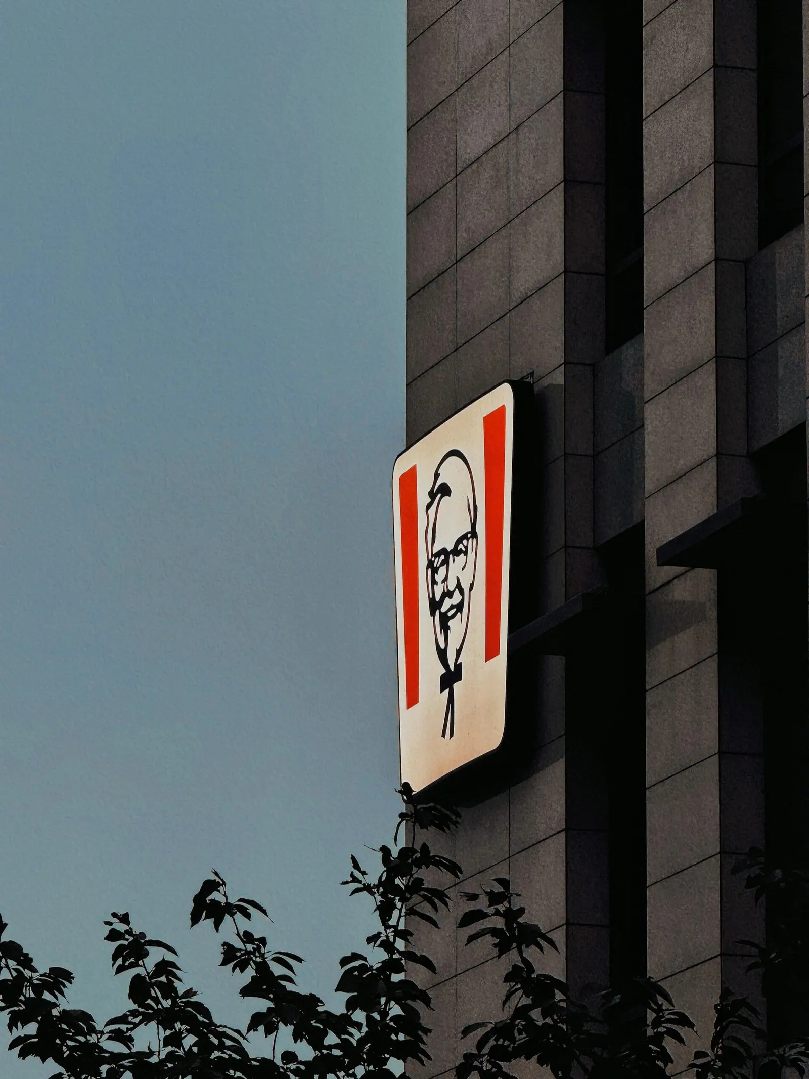 A lit KFC sign with the Colonel Sanders face on the side of a stone building at dusk