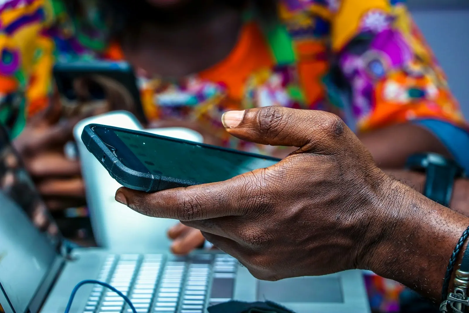 A man's hand holding a black Android smartphone over a laptop keyboard in Lagos, with a woman in a bright print shirt behind
