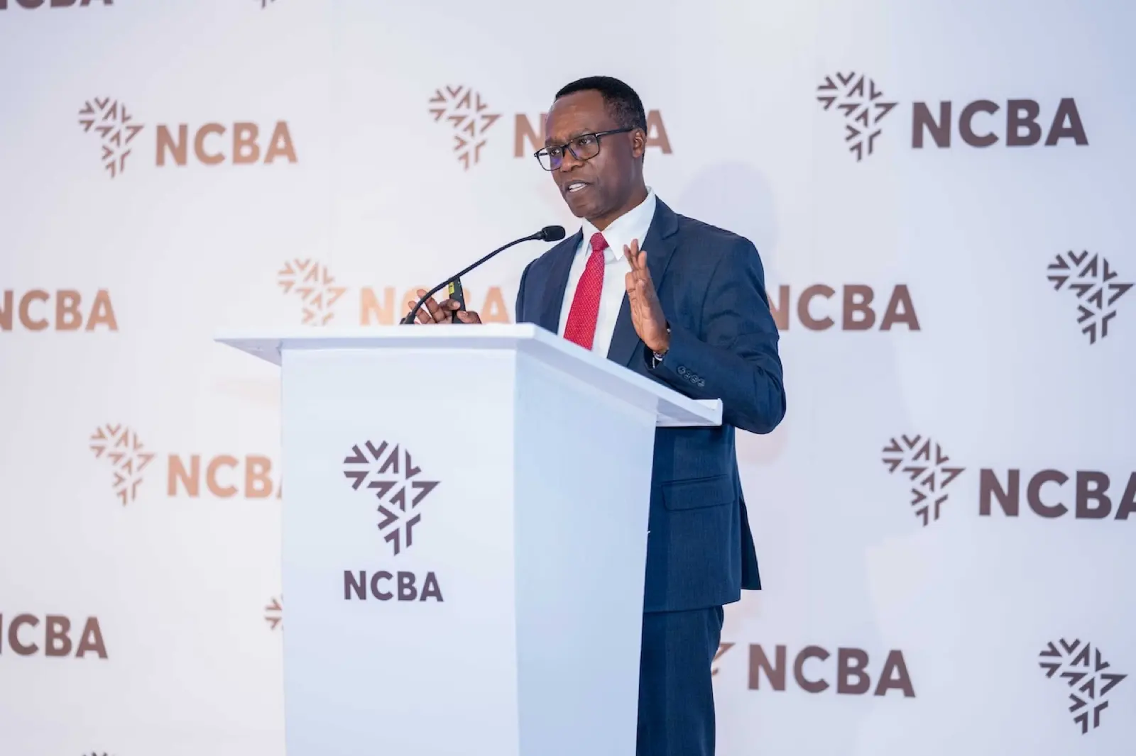 NCBA Group chief executive John Gachora speaks at a lectern in front of an NCBA-branded backdrop