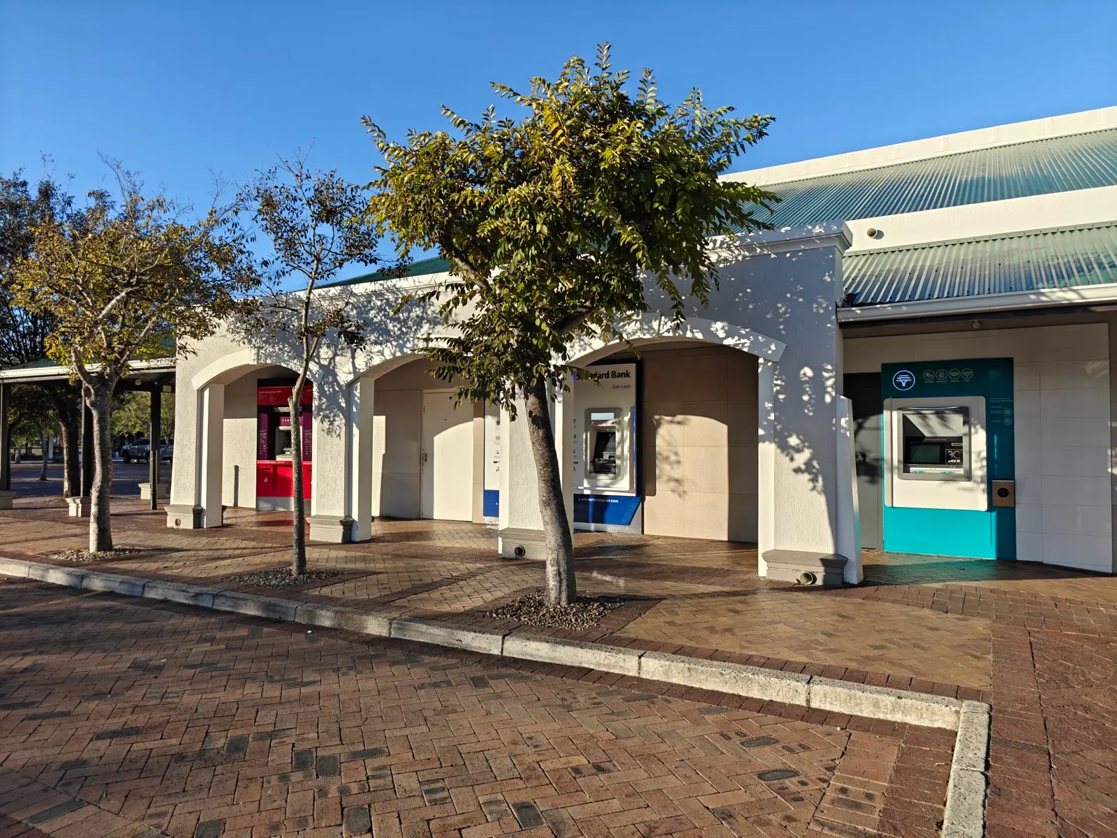 Three bank-branded ATMs side by side under arches at a shopping centre in Constantia, Cape Town, in red, blue and teal